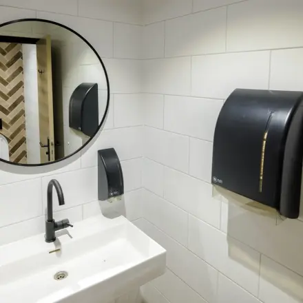 Moderne toiletruimte in een kantoorpand aan de Hogehilweg 24, Amsterdam Zuid-Oost, met witte betegelde muren, een ronde spiegel en een wastafel.