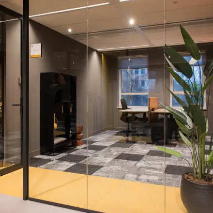 Moderne kantoorruimte huren in Amsterdam Zuid-Oost met glazen wanden en grijze vloertegels. Voorzien van een bureau, stoel en grote kamerplant, geschikt voor professionele activiteiten.