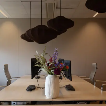 Moderne kantoorruimte huren in Apeldoorn met stijlvolle vergadertafel, comfortabele bureaustoelen en opvallende hanglampen. Groot bloemstuk op tafel, geschikt voor creatieve bijeenkomsten.