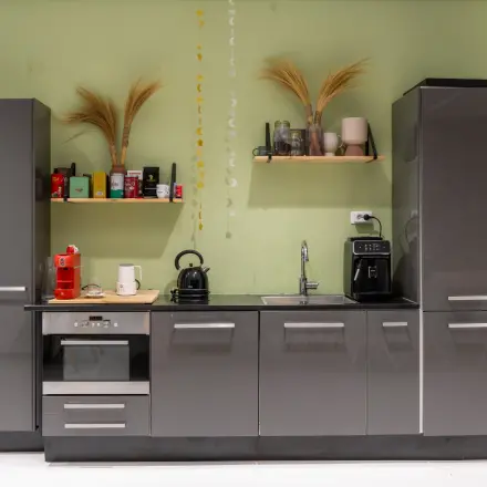 Gemeubileerde pantry van een kantoorpand met moderne grijze kastjes, een koffiezetapparaat en decoratieve elementen. Ideaal voor wie kantoorruimte wilt huren in Baarn.