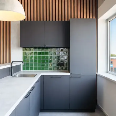 Moderne keuken in kantoorpand op Tolnasingel 1-3, Bodegraven, met grijskleurige kasten en groen betegelde achterwand, verlichting en groot raam. Geschikt voor korte pauzes in een open floorplan kantoor.