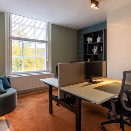 Moderne kantoorruimte te huur in Breda Centrum-Station met twee werkplekken, bureaustoelen, en een leeshoek met fauteuil bij het raam.