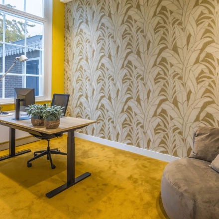 Kantoorruimte huren met geel tapijt en opvallende printbehang op de muur, bureau met stoelen en plantdecoratie, raam biedt natuurlijk licht in het pand aan de Catharinastraat 16 in Breda Centrum-Station.