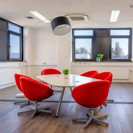 Vergaderruimte in kantoorpand Druivenstraat 25-31, Breda Oost, met rode stoelen rond een ovale tafel. Veel lichtinval door grote ramen. Geschikt voor kantoorruimte huren.