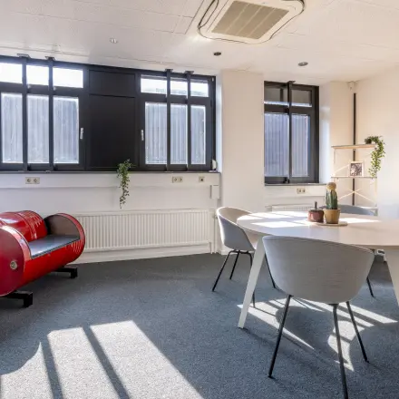 Lichte kantoorruimte met grote ramen, ronde tafel met stoelen en een decoratieve open haard in kantoorpand op Druivenstraat 25-31, Breda Oost. Ideaal voor kantoorruimte huren.