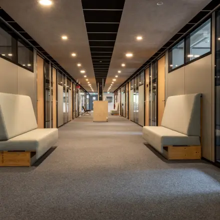 Lichte gang in modern kantoorpand op Delftechpark 9, Delft, met glazen kantoorruimten aan beide zijden en grijze vloertapijten. Benches langs de muren bieden zitmogelijkheden. Ideaal voor kantoorruimte huren.