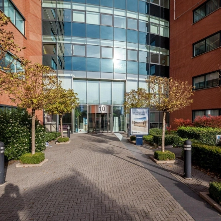 Toegang tot kantoorgebouw aan de Olof Palmestraat 10 in Delft, met moderne glazen entree omgeven door bakstenen gevel en groenvoorzieningen. Ideale locatie voor kantoorruimte huren.