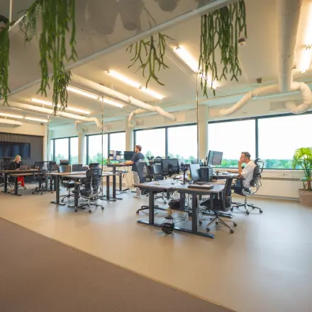 Modern kantoor op Hambakenwetering 1, Den Bosch, met open werkruimtes waar mensen zakelijk aan bureaus werken. Ideaal voor kantoorruimte huren, met ruime ramen en natuurlijke elementen.