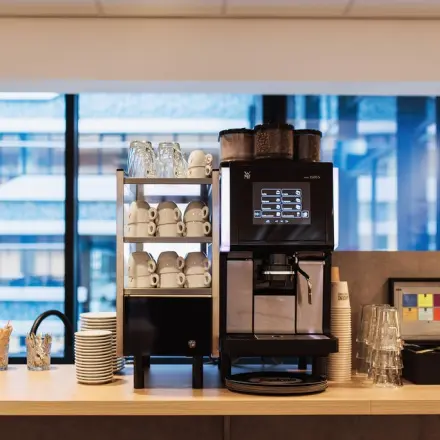 Moderne koffiehoek in een kantoorpand aan Anna van Buerenplein 41, Den Haag Centrum, met een koffiemachine, bekers en glazen tegen een glazen achtergrond.