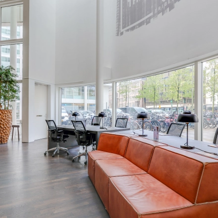 Moderne kantoorruimte met grote ramen, voorzien van een lange tafel met stoelen, leren bank en veel natuurlijke lichtinval op Kalvermarkt 53, Den Haag Centrum.