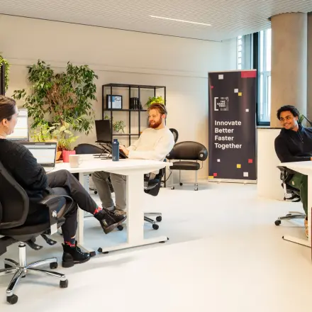 Zakelijke omgeving op Waldopstraat 5, Den Haag Centrum, met mensen in gesprek in een moderne kantoorruimte. De ruimte is voorzien van bureaus en enkele planten.