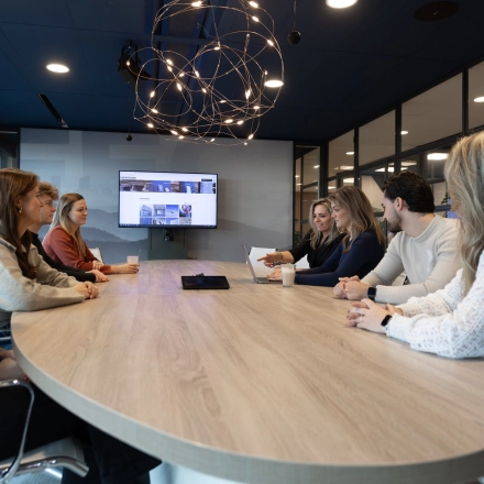 Zakelijk overleg in een modern kantoorpand op Flight Forum 120-159, Eindhoven Airport. Zes personen zijn in gesprek rond een houten tafel, met een groot scherm op de achtergrond. Ideaal voor kantoorruimte huren.
