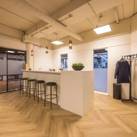 Moderne kantoorruimte huren bij Eindhoven Airport, Luchthavenweg 81. Het interieur omvat een stijlvolle bar met krukken, een vloerkleed in houtlook en een kapstok, ideaal voor zakelijke bijeenkomsten.