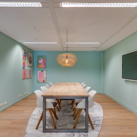 Vergaderruimte met lange houten tafel en moderne stoelen op Stadhuisplein 10 in Eindhoven Centrum. Kleurrijke kunstwerken aan de muur en een grote monitor maken deze plek geschikt voor kantoorruimte huren.