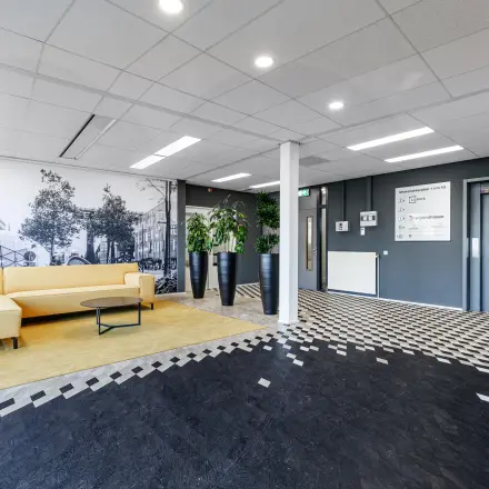 Een moderne kantoorlobby met een beige bank, planten en muurdecoraties in een gebouw op Meerenakkerplein 1-10, Eindhoven De Hurk. Ideaal voor kantoorruimte huren in dit kantoorpand.