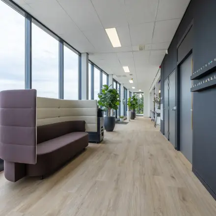 Moderne kantoorruimte huren in Eindhoven De Hurk met een smalle gang, voorzien van glazen wanden, houten vloer en zitmeubilair. Veel lichtinval en planten sieren het strakke interieur.