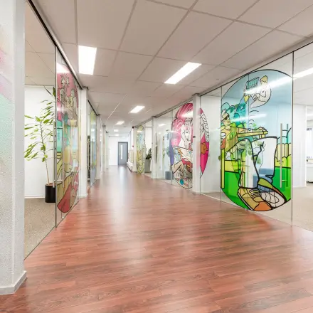 Kleurrijk kantoorpand op Meerenakkerplein 11-20 in Eindhoven De Hurk, met glaspanelen met kunstwerken langs de gang. Ideaal voor wie kantoorruimte wil huren met een inspirerende uitstraling.