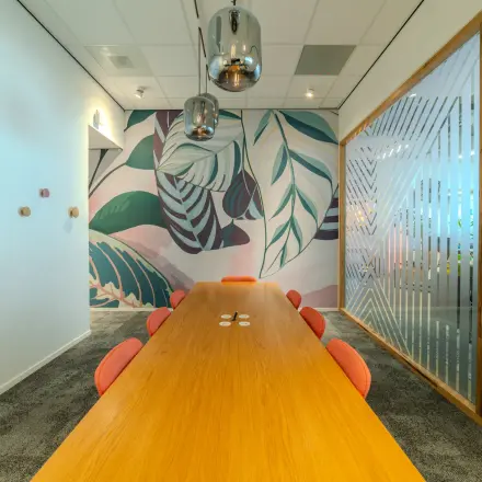Moderne vergaderruimte op Torenallee 20, Eindhoven Strijp-S, met een lange houten tafel, kleurrijke stoelen en een opvallende muurschildering. Ideale plek om kantoorruimte te huren.