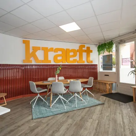 Moderne en ingerichte kantoorruimte aan de Boddenkampsingel 95 in Enschede met tafels, stoelen en planten. Grote oranje letters op de achtergrond en een comfortabele werkplek voor het huren van kantoren.