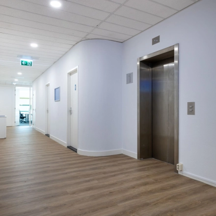 Een lichte gang in een kantoorpand met een houten vloer, witte muren en een moderne lift. De ruimte is goed verlicht en leidt naar verschillende kantoorruimtes.