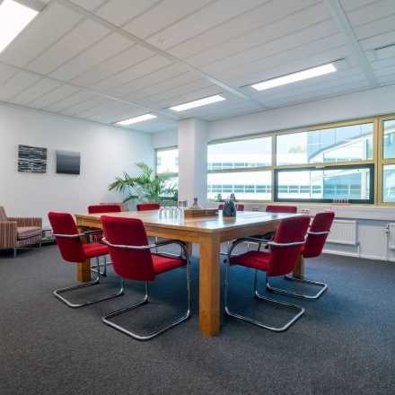 Ruime vergaderkamer in kantoorpand aan Groningenweg 8 8, Gouda, met rode stoelen rondom een houten tafel, grote ramen en kamersplanten. Ideaal voor kantoorruimte huren.