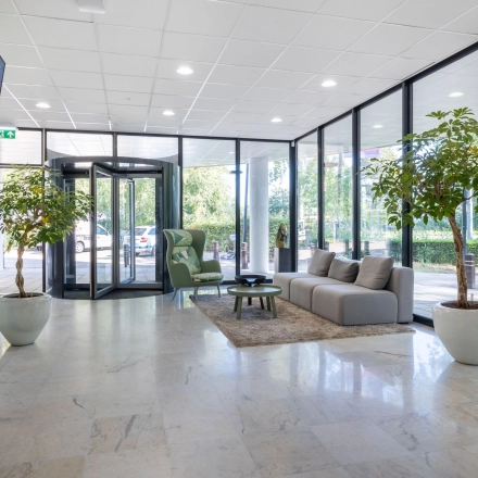 Lichte entree van een kantoorpand in Gouda met glazen deuren, een comfortabele bank, en planten. Ideaal voor kantoorruimte huren.