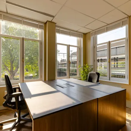 Lichte kantoorruimte te huur op Kennemerplein 20, met grote ramen, uitzicht op Haarlem Centraal Station en voorzien van bureaus en stoelen.