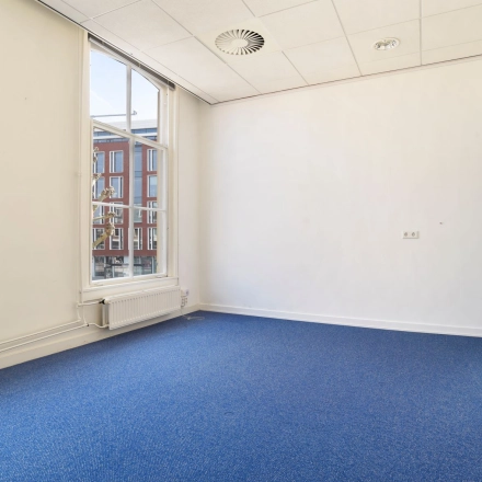 Leeg kantoorpand op Houtplein 22, Haarlem Zuid met blauwe vloerbedekking en grote ramen die daglicht binnenlaten, ideaal voor een creatieve inrichting voor wie kantoorruimte wil huren.