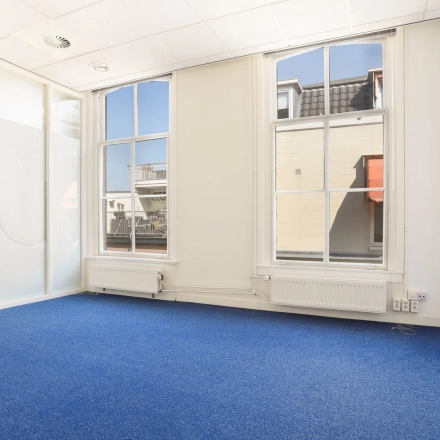 Lichte kantoorruimte op Houtplein 22, Haarlem Zuid, met grote ramen en blauwe vloerbedekking, geschikt voor zakelijke activiteiten. Ideaal voor het huren van een moderne werkplek.