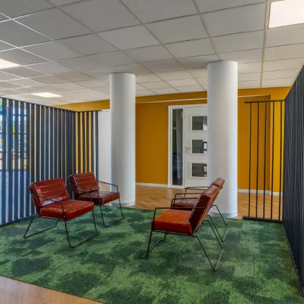 Moderne kantoorruimte met kleurrijke muren en comfortabele zithoek, ideaal voor informele vergaderingen of pauzes. Glaswand zorgt voor veel licht en een uitnodigende sfeer om te werken.