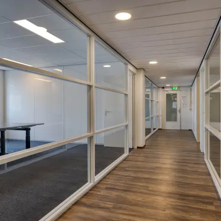 Glazen wanden verdelen de kantoorruimte op Franciscusweg 219 in Hilversum met houten vloeren en zicht op ramen langs de gang.