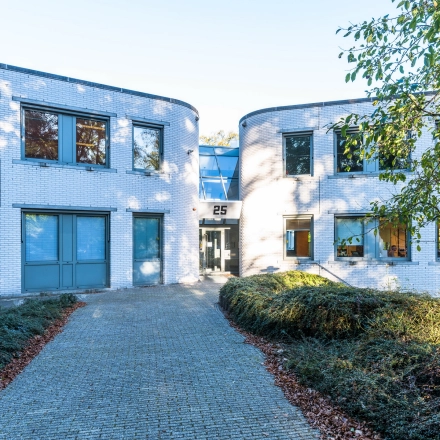 Uitzicht op de buitenkant van het kantoorpand op Mozartlaan 25 in Hilversum, geschikt voor kantoorruimte huren. Het entreepad leidt naar een moderne glazen ingang die omringd is door witte bakstenen muren.