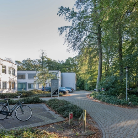 Kantoorpand aan de Mozartlaan 25 in Hilversum, omgeven door bosrijke omgeving. Parkeerplaats met fietsen en auto's zichtbaar.