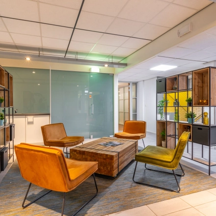 Moderne kantoorlounge in Hilversum met houten rekken, kleurrijke stoelen en een glazen wand, ideaal voor ontspanning tijdens de werkdag. Kantoorruimte huren met verfijnde inrichting.