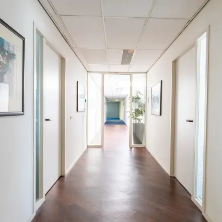 Heldere gang in een kantoorpand op Sandtlaan 36, Katwijk, met houten vloer en kunst aan de muur. Ideaal voor bedrijven die kantoorruimte willen huren met ruimte voor creativiteit en productiviteit.