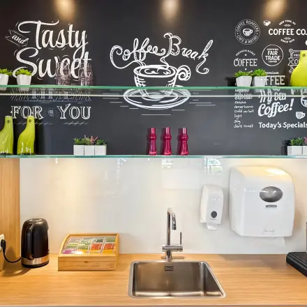 Koffiehoek in een kantoorpand met glaspanelen, muur met koffie-gerelateerde teksten en planken met decoratieve elementen. Kleine wastafel en koffiemachine aanwezig.
