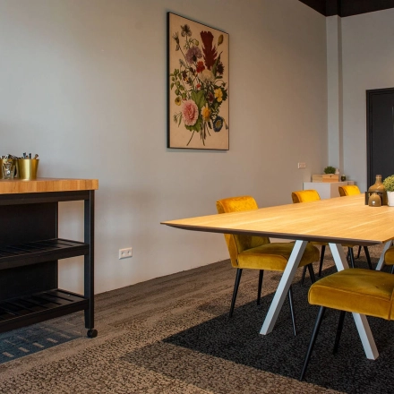 Moderne kantoorruimte in Meppel met vergadertafel en felgekleurde stoelen, ingericht om professioneel te huren. Er is een koffiezetapparaat en een beeldscherm aan de muur.