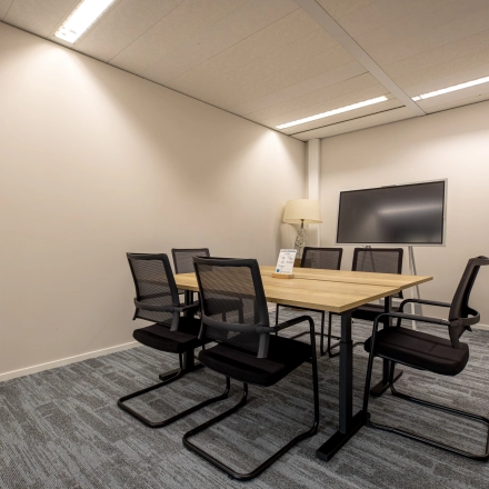 Moderne vergaderruimte in kantoorpand op Gooimeer 1, Naarden, met zes stoelen rond een houten tafel en een groot beeldscherm aan de muur. Ideaal voor bedrijven die kantoorruimte willen huren.