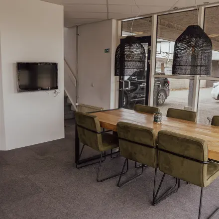 Kantoorruimte huren in Oosterhout Nb, Abdis van Thornstraat 74. Modern kantoorpandinterieur met houten tafel en groene stoelen, tegen een glazen wand met uitzicht op buiten. Neutrale tinten en stijlvolle verlichting.