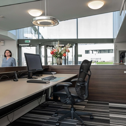 Receptieruimte in een kantoorpand aan Boven de Wolfskuil 3, Roermond, met balie, computerscherm en stoelen. Een persoon staat binnen te kijken vanuit de ingang.