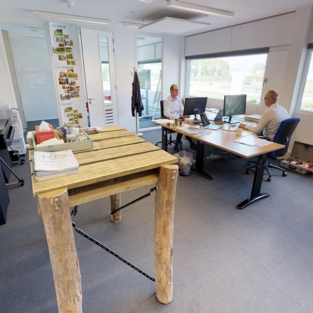 Modern kantoorpand in Roermond met lichte werkplekken, ideaal voor kantoorruimte huren. Twee mensen overleggen aan bureaus. Ruime ramen zorgen voor veel natuurlijk licht.