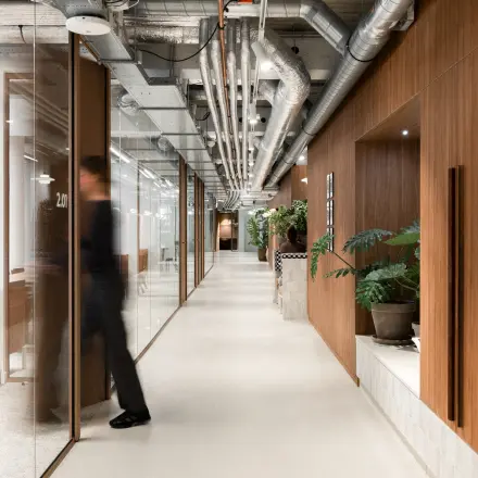 Moderne kantoorruimte te huur op Stadhuisplein 9-23 in Rotterdam Centrum, met glazen wanden, leidingen aan het plafond en groene planten. Een persoon loopt door de gang richting een kantoor.