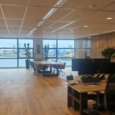 Ruime kantoorruimte met houten vloer en uitzicht op de haven. De ruimte heeft werkplekken, planten en biedt diverse zitmogelijkheden. Ideaal voor bedrijven die kantoorruimte willen huren in Rotterdam Waalhaven.