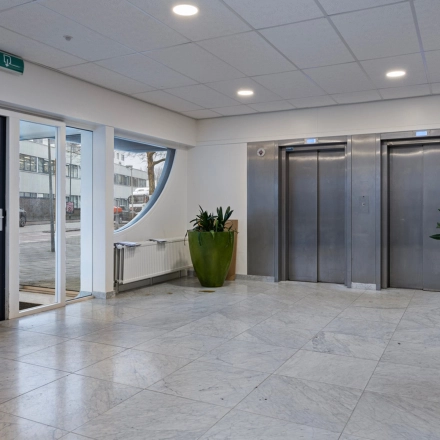 De entree van het kantoorpand aan de Brauwweg 52-66 in Schiedam. Moderne stenen vloer, groene planten naast liften, en ruime toegangsdeur benadrukken de functionele designstijl.