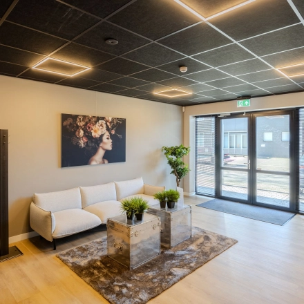 Moderne ontvangstruimte in een kantoorpand met een stijlvolle witte bank, glazen salontafel en kunst aan de muur. Gezellige verlichting en groene planten verfraaien de ruimte. Ideaal voor kantoorruimte huren.