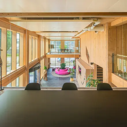 Moderne kantoorruimte met houten interieur en grote ramen, geeft uitzicht op een open zicht van meerdere verdiepingen met kleurrijke meubels en planten, geschikt voor diverse werkactiviteiten.