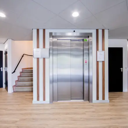 Ingang van een kantoorpand in Woerden op Pelmolenlaan 16, met een lift en trap. Sfeervol verlicht met houten accenten. Kantoorruimte huren mogelijk.