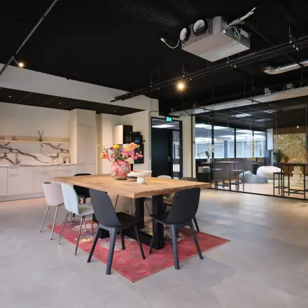 Stijlvolle kantoorruimte huren in Woerden, Vijzelmolenlaan 9. Moderne inrichting met een grote tafel, kleurrijke bloemen, en een ontspannende zithoek. Perfect voor zakelijke bijeenkomsten en informele vergaderingen.