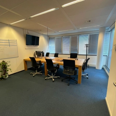 Vergaderkamer in kantoorpand met grote houten tafel, zes bureaustoelen, en aanwezigheidsmonitoren aan de muur op Baron de Coubertinlaan 35, Zoetermeer. Ideaal voor kantoorruimte huren.