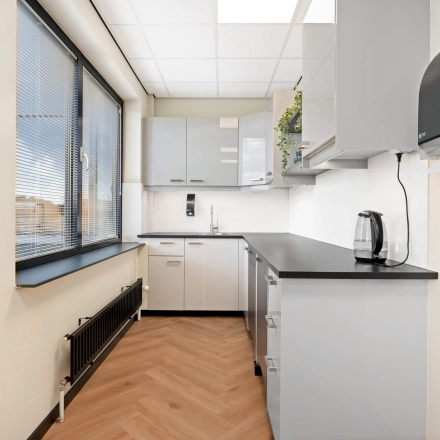 Moderne pantry in kantoorpand op Stationsplein 4, Zwijndrecht, voorzien van strakke keukenkasten, een waterkoker en een groot raam. Perfecte locatie om kantoorruimte te huren. Functioneel en uitnodigend ontwerp.